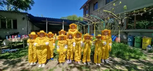 Дванайсет деца в жълти пчеларски костюми с шапки с воал стоят в редица в двора на хижа под синьо небе.
