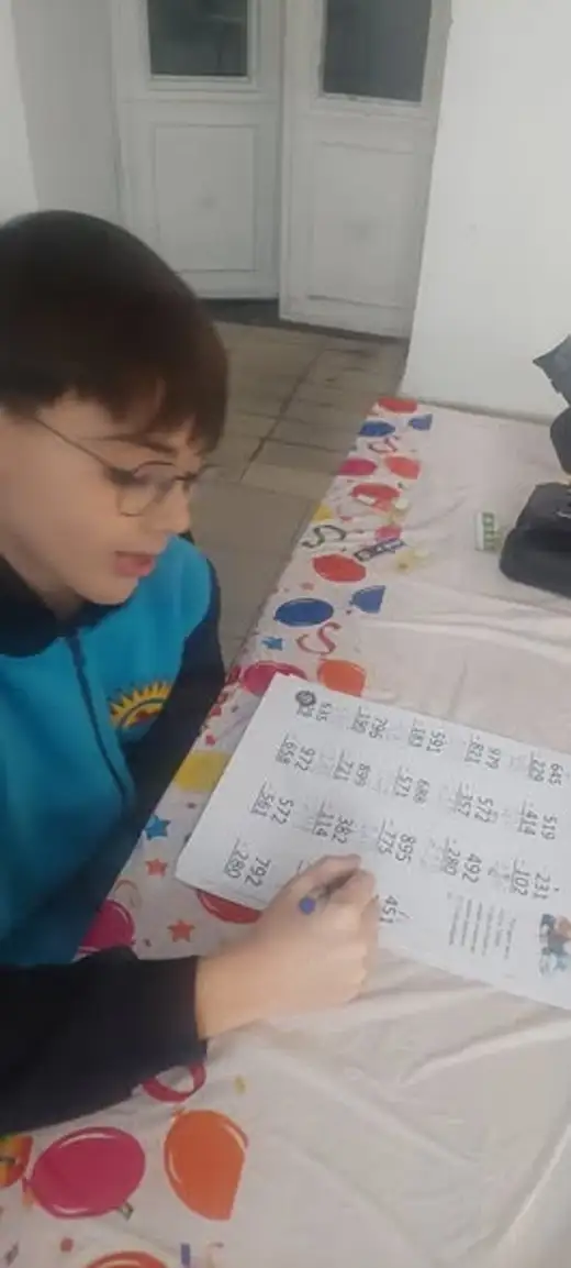 Момче пише в тетрадка на маса с цветна покривка.