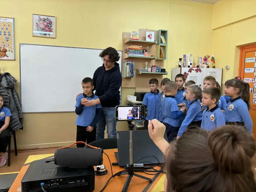 Ученици в класна стая около телефон на статив, който записва.