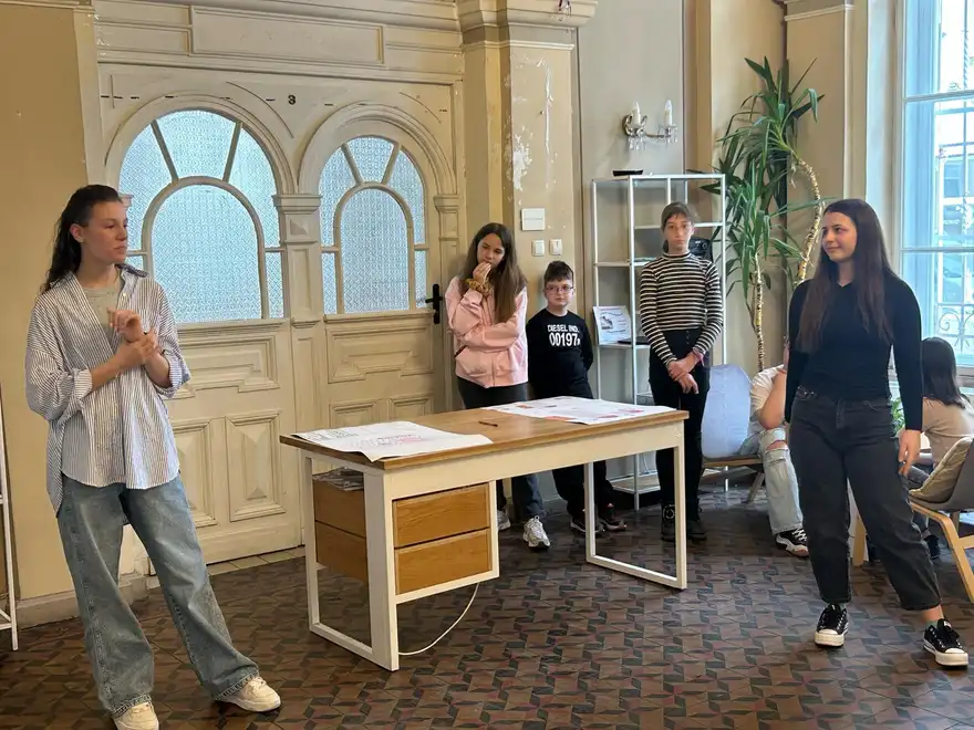 Момиче говори пред група деца в зала с висок таван; на бюрото до нея са разгънати листове с рисунки