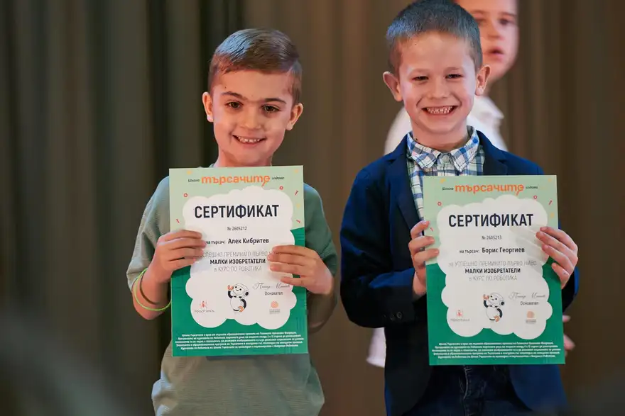 Две момчета вдигат зелено-бели сертификати с надпис „Сертификат“ пред тъмна завеса.