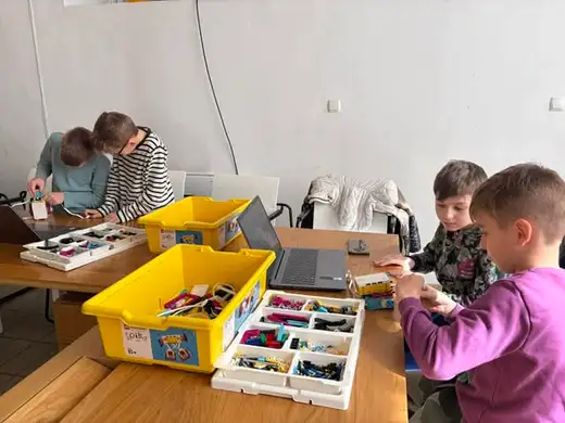 Деца се навеждат над маса с жълта кутия с детайли, отзад жена подрежда материали.