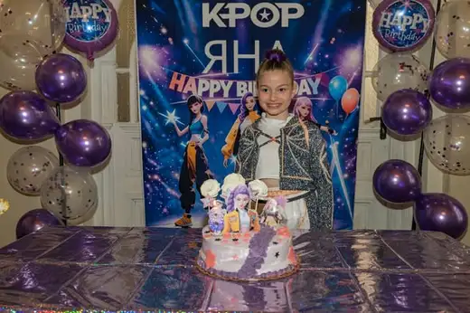 Момиче стои пред пано с надпис K-POP, между лилави и прозрачни балони, а на масата пред него торта с фигурки.