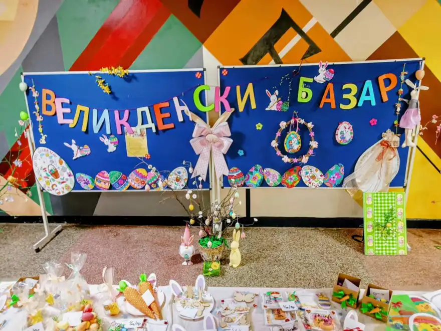 Табла с надпис „Великденски базар“, украсени с хартиени яйца и зайчета, а пред тях маса със сувенири